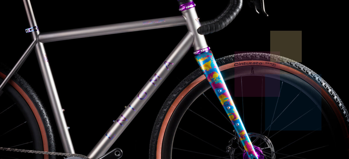 Side view of Enigma titanium gravel bike highlighting the custom-painted fork in blue, yellow, and magenta, purple Chris King components, and Pirelli Cinturato tyres against a black background.