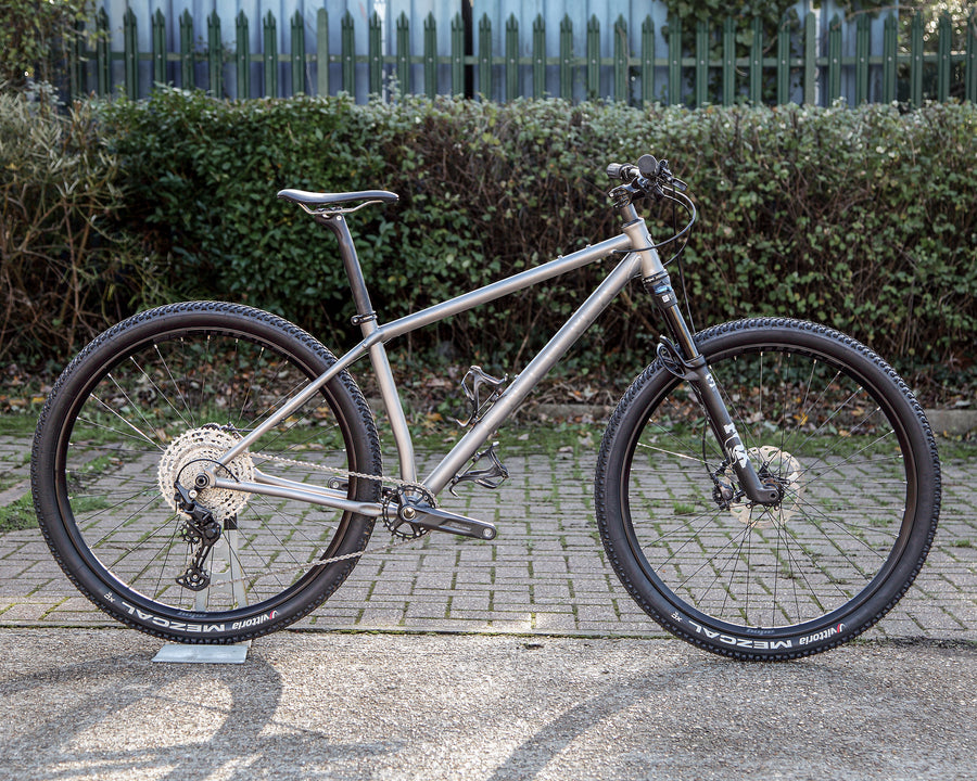 Enigma Explor Medium titanium flat-bar adventure bike, side-on view showing satin bead frame with brushed logos, Shimano Deore XT drivetrain, Fox 32 suspension fork and Hope Fortus wheels.