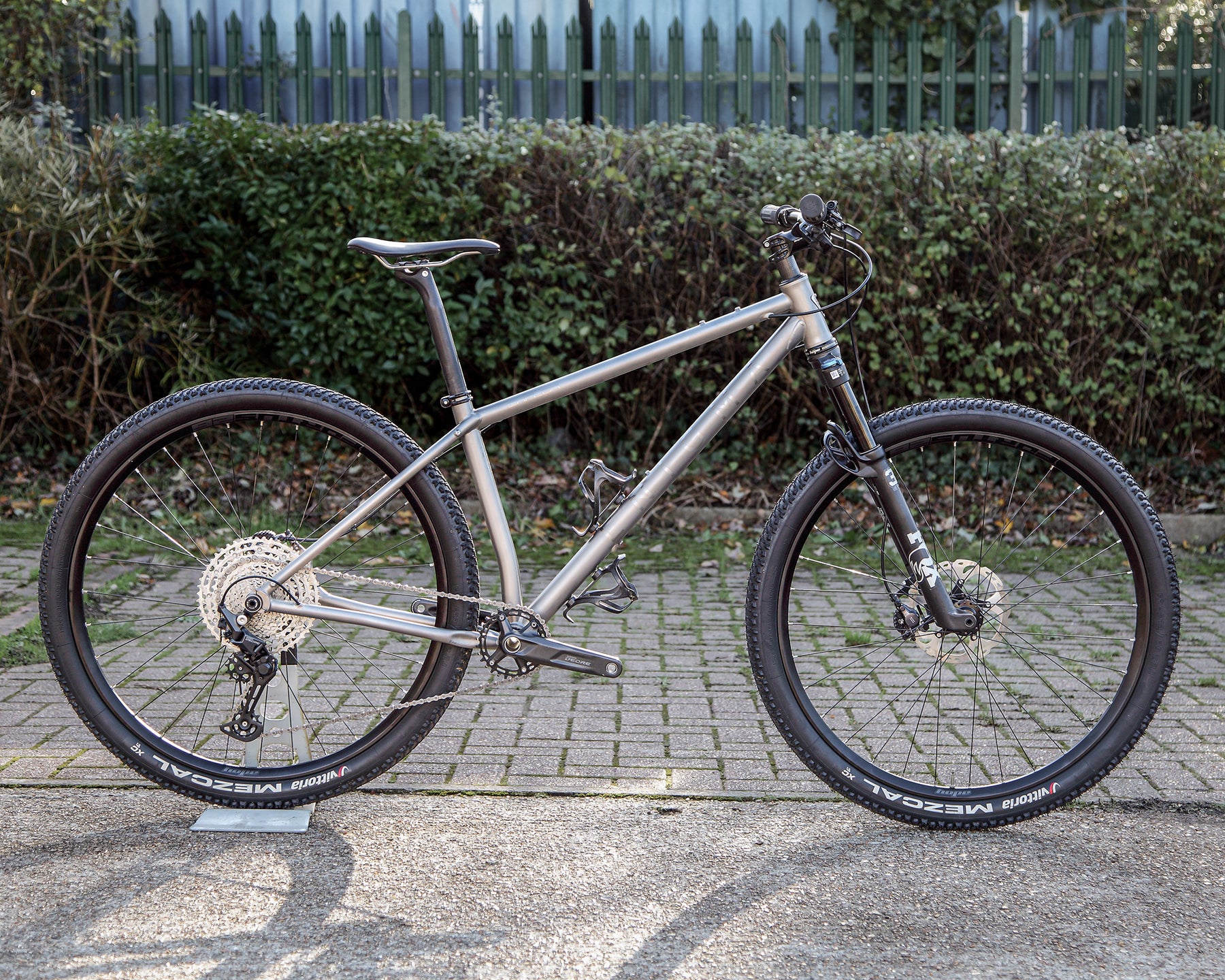 Enigma Explor Medium titanium flat-bar adventure bike, side-on view showing satin bead frame with brushed logos, Shimano Deore XT drivetrain, Fox 32 suspension fork and Hope Fortus wheels.