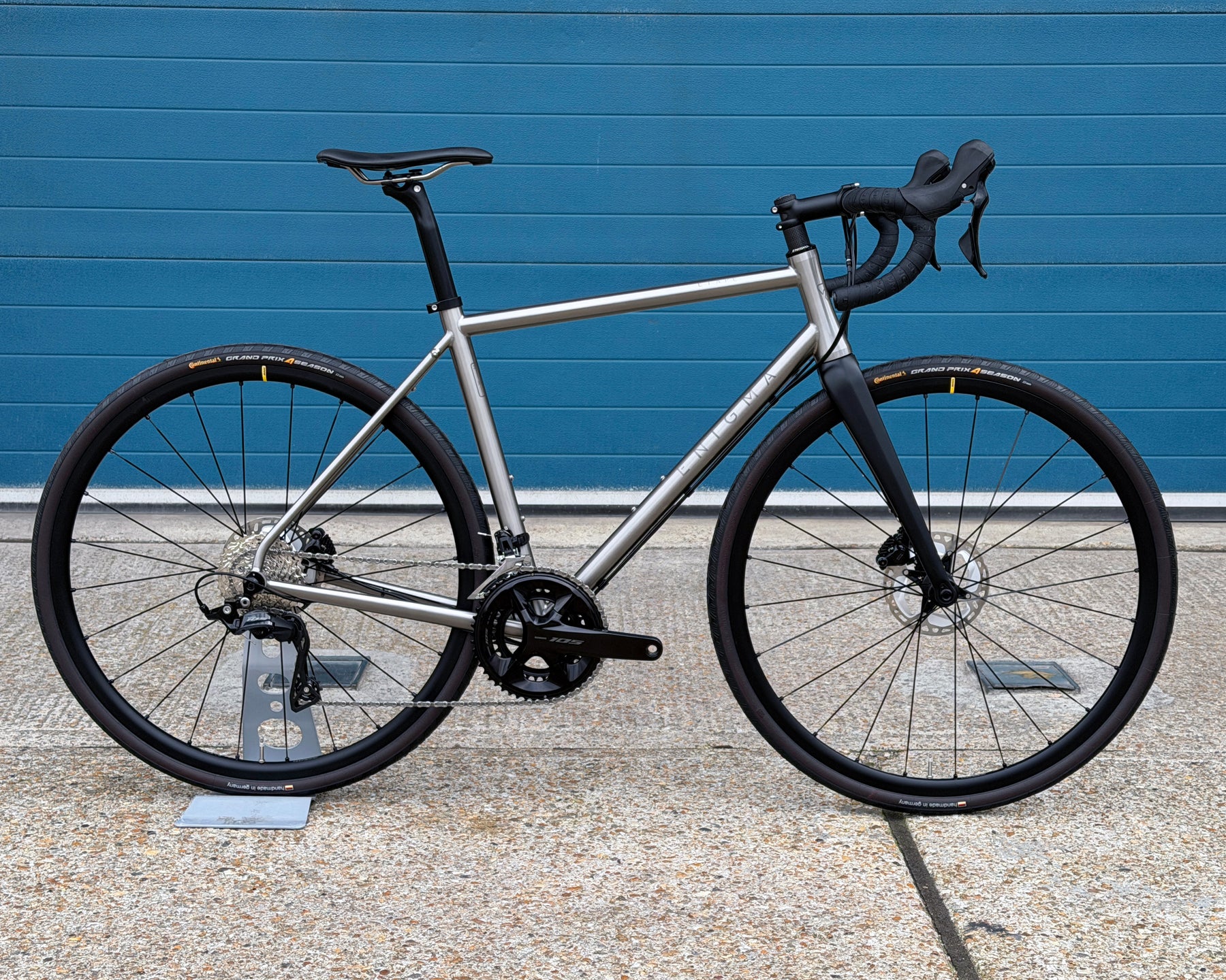 Titanium Enigma bicycle with black tyres against a blue wall